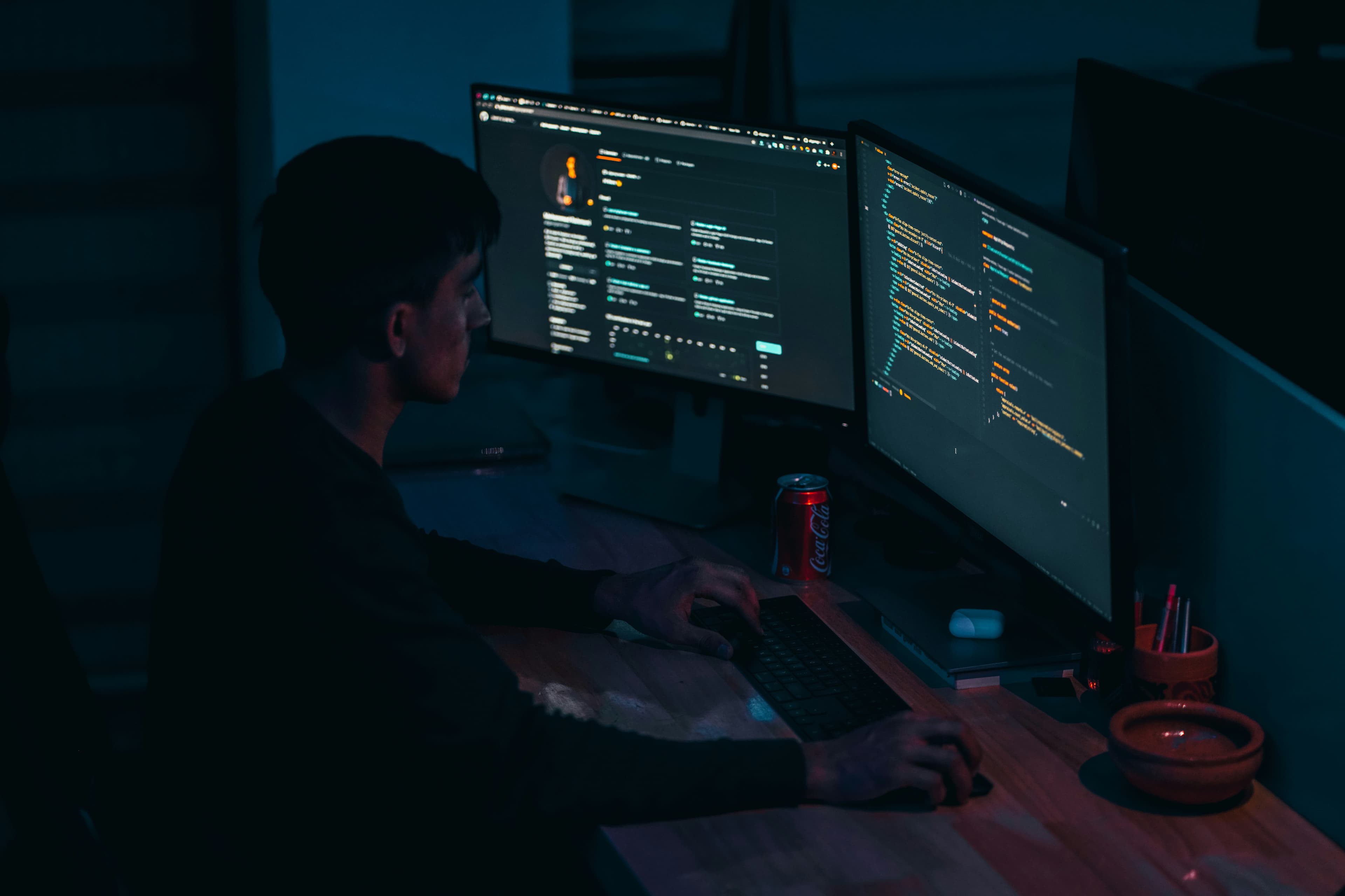 Developer at dual monitors in dark office, blue ambient lighting, focused coding environment